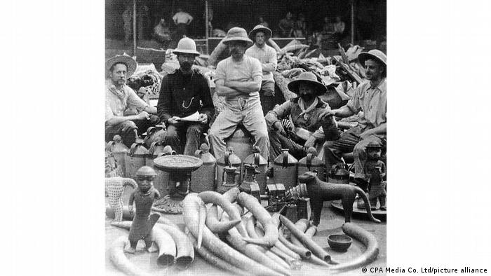 Männer mit Hüten sitzen um Elfenbeinstoßzähne und Bronzestatuen